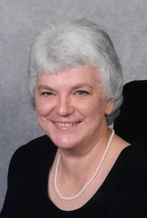 Portrait of a smiling woman with short white hair, wearing a black top and pearl necklace.