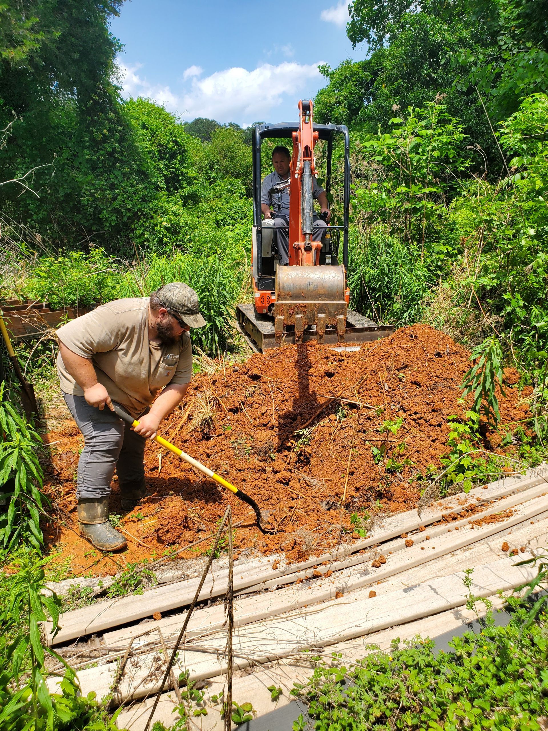 a man is digging a hole in the dirt with a shovel while a man is driving a small excavator