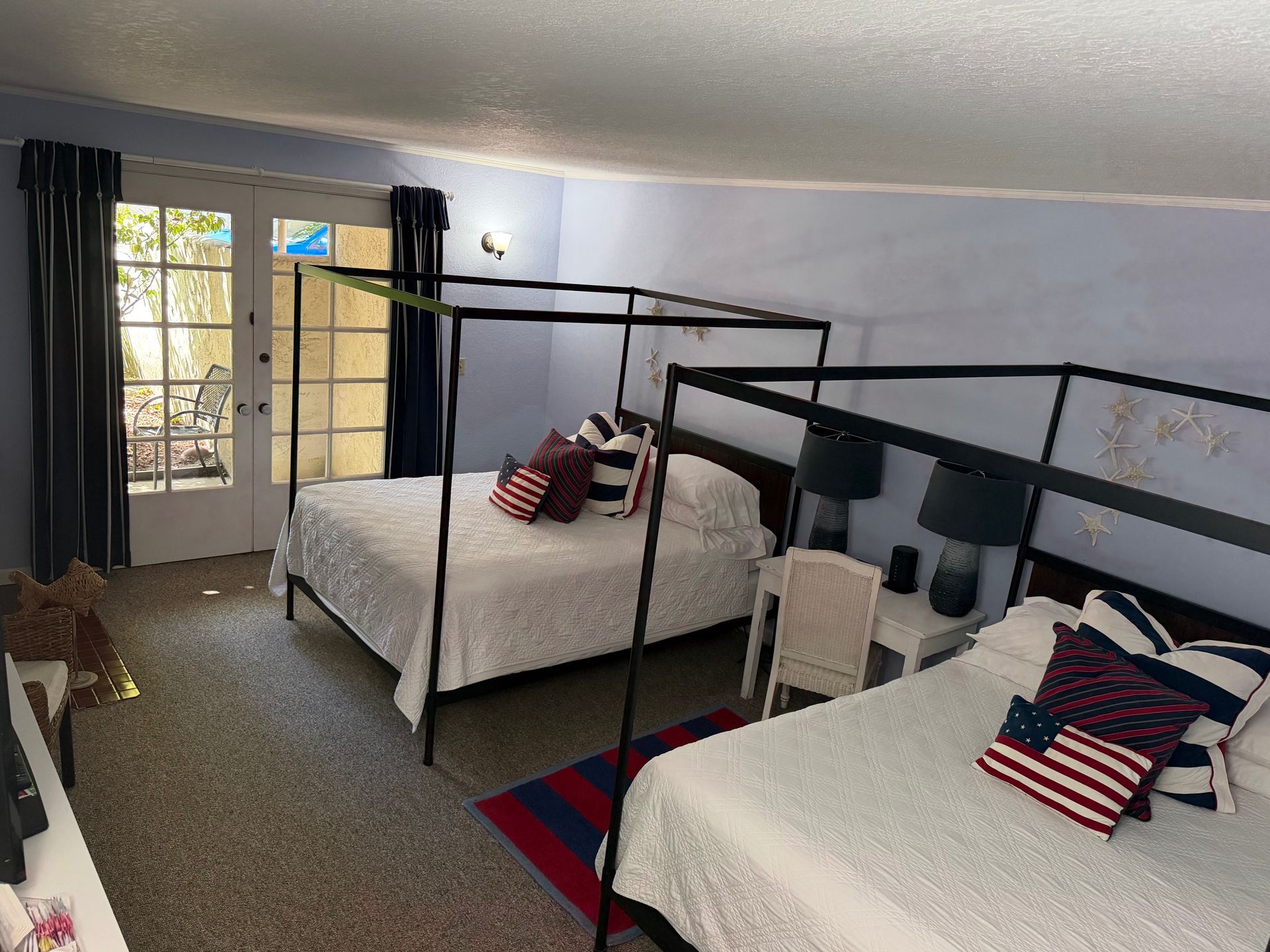 Two beds in a room with patriotic pillows, black canopy frames, a rug, and french doors.