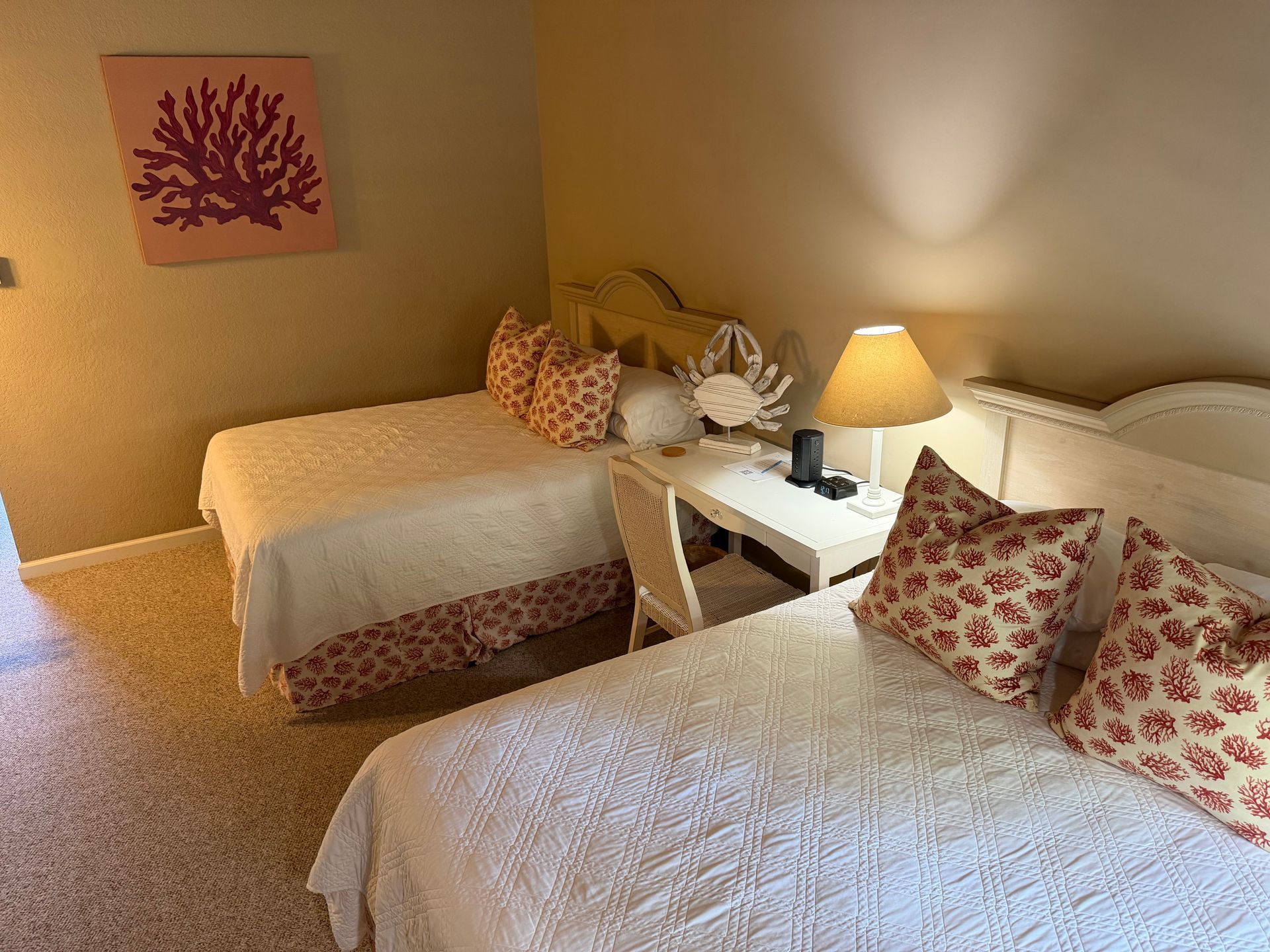 Two beds in a room, white quilts, patterned pillows, coral artwork, beige walls, small desk.