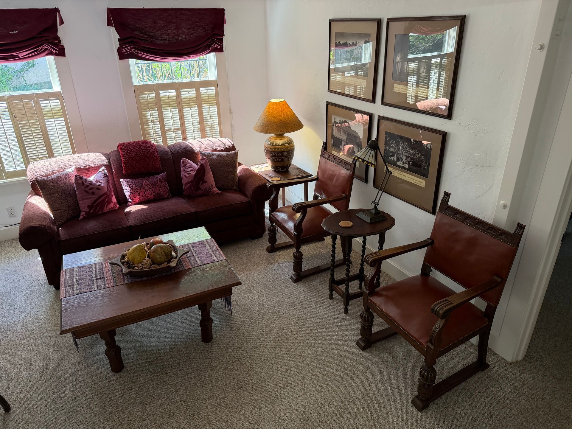 Cozy living room with burgundy couch, coffee table, and chairs. Walls adorned with framed art, soft light.