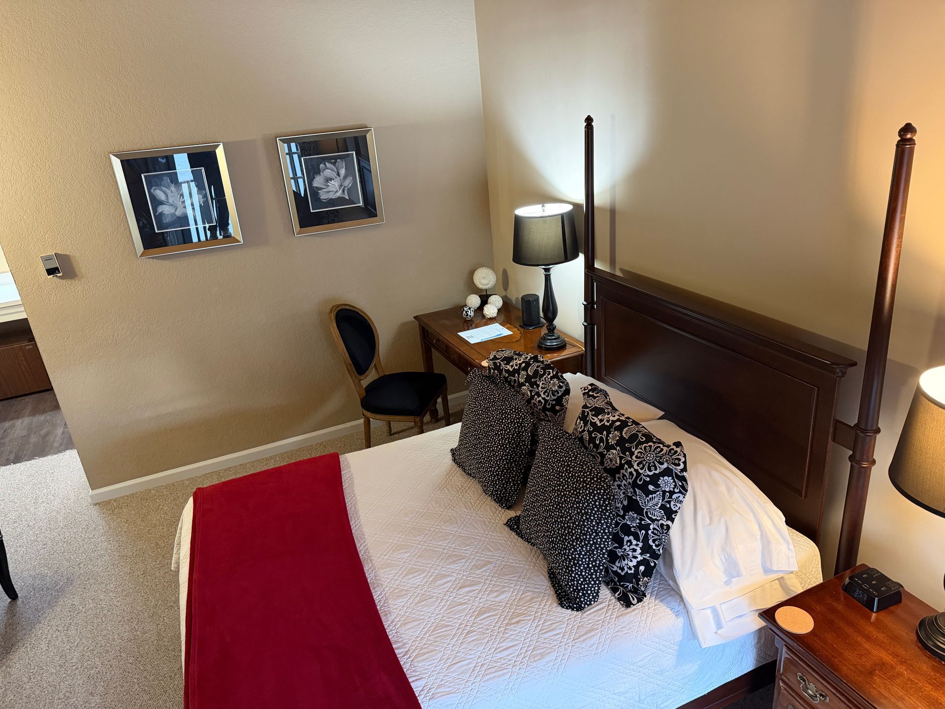 Bedroom with a made bed, two framed pictures on the wall, and a bedside table with a lamp.