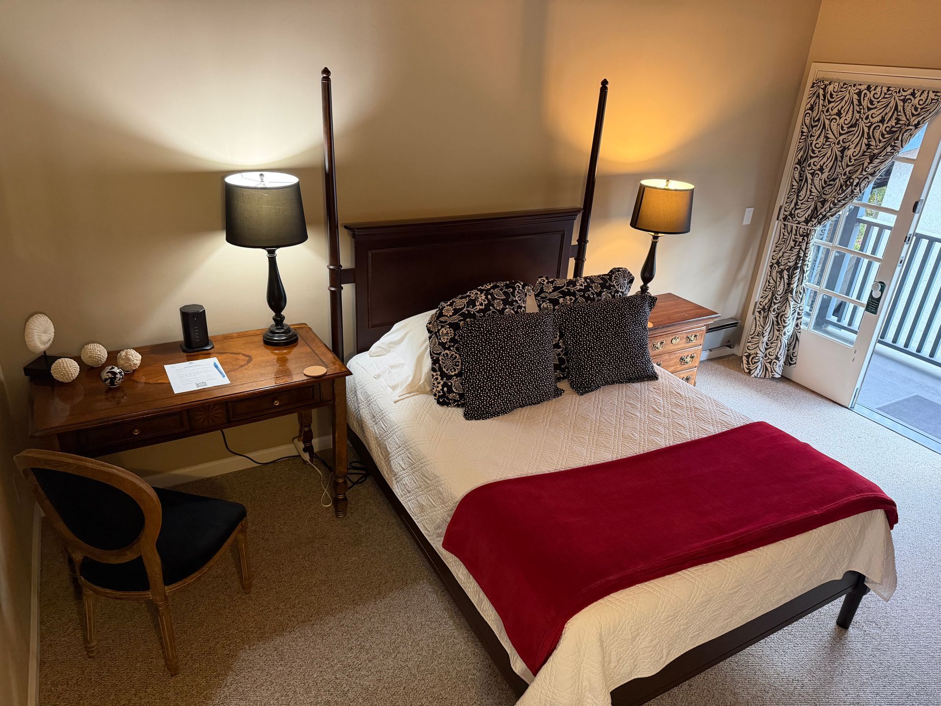Bedroom with a dark wood four-poster bed, desk, chair, and door to a balcony. Red throw on the bed.