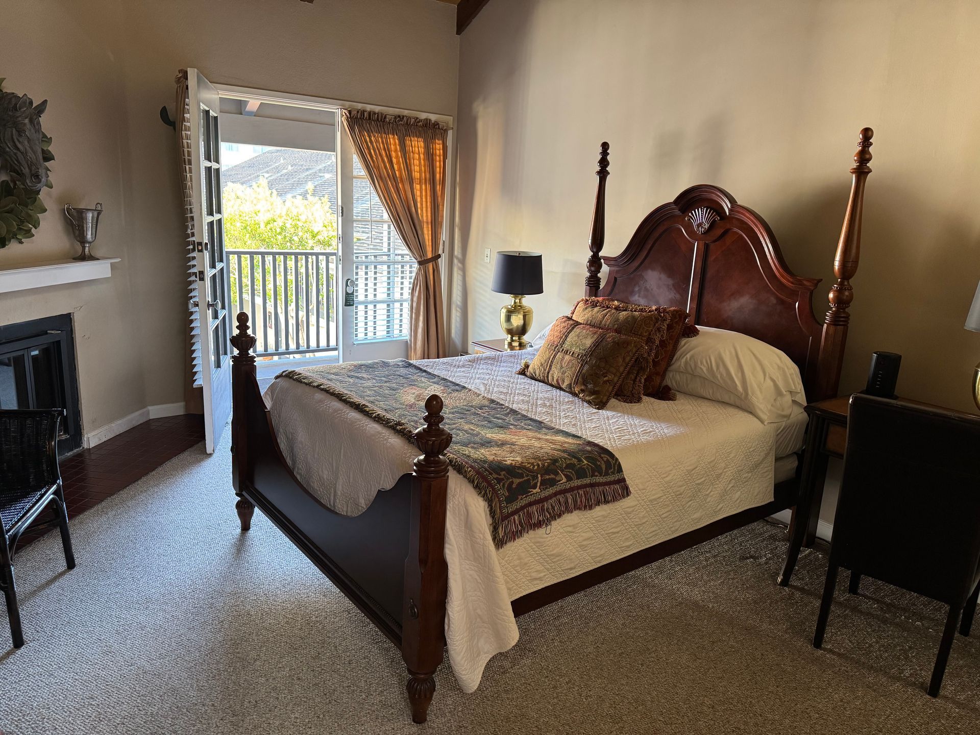 Bedroom with a four-poster bed, open balcony doors, and a fireplace. Beige walls, carpet; dark wood furniture.