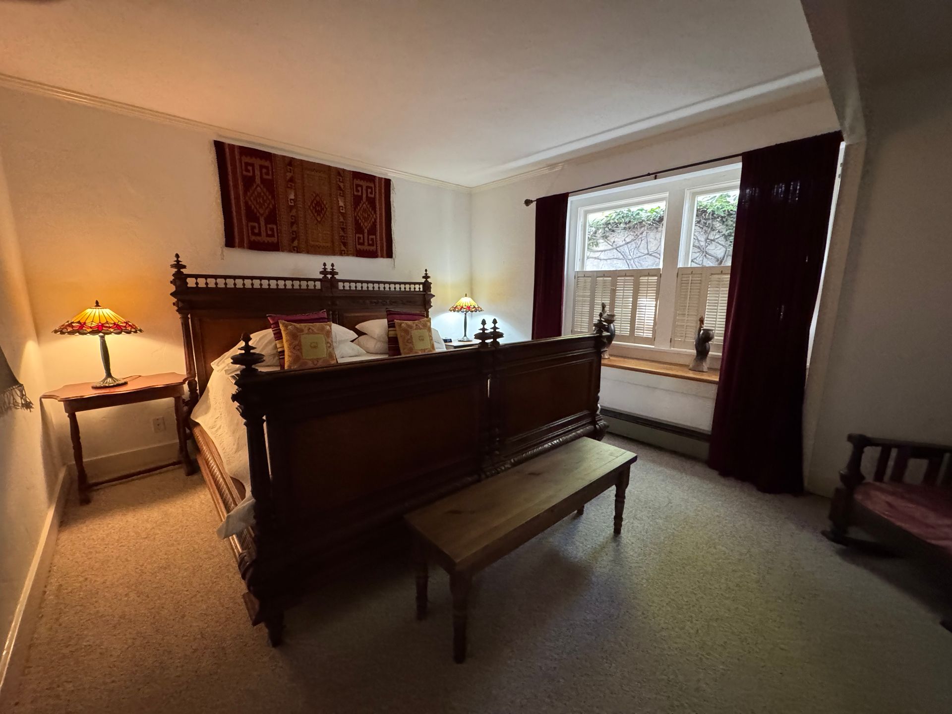 Bedroom with dark wooden bed, bench, and side table. Red and cream textiles, window with shutters.