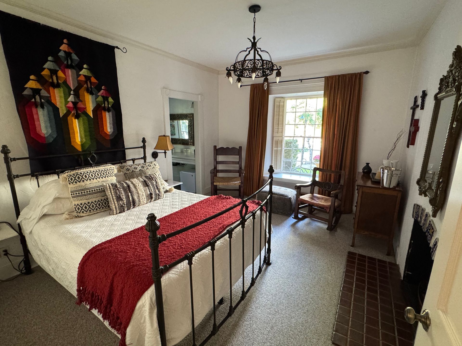 Bedroom with iron bed, red blanket, rug, tapestry, fireplace, and window with curtains.