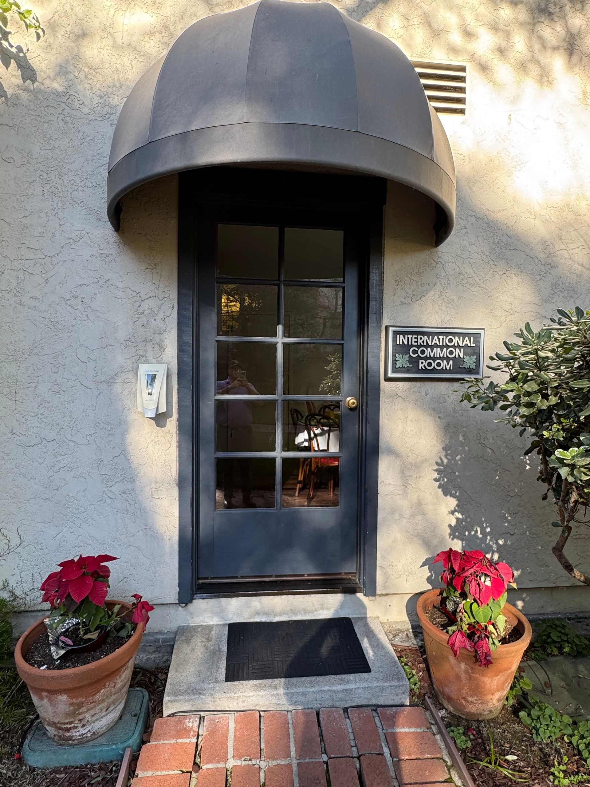 Entrance to a building with a black door, awning, and nameplate. Two potted plants flank the entrance.