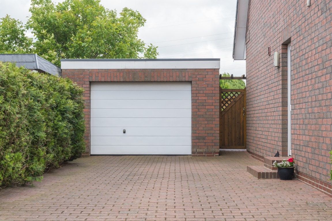 Driveway leading to a white garage door beside brick house walls and green hedges.