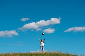 Person standing on grass, reaching up toward a ball in a blue sky with clouds
