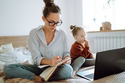Woman writing in a notebook beside a child using a laptop on a bed in a bright bedroom