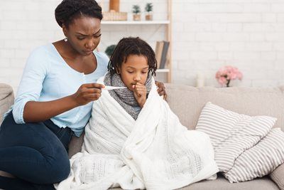 Adult caring for a child wrapped in a blanket on a couch, with a tissue in hand.