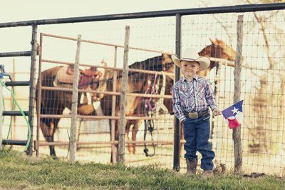 Child in cowboy hat stands by a fence with horses, holding a small Texas flag.