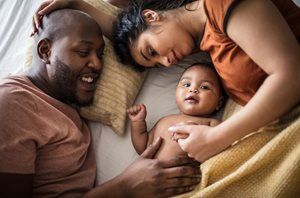 Family cuddling with a baby on a bed, smiling under a beige blanket