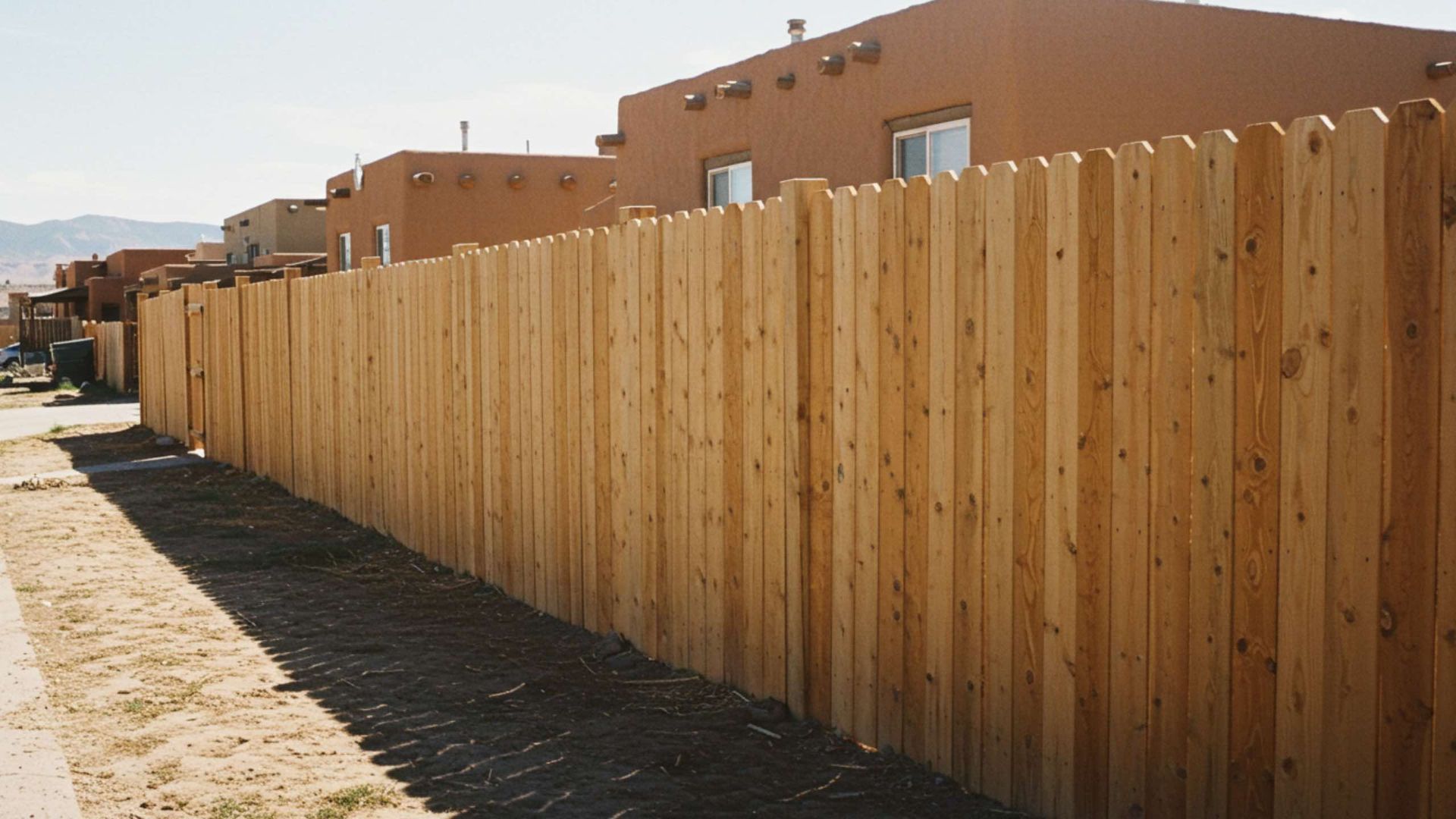Wooden fence along a street with tan buildings in the background.