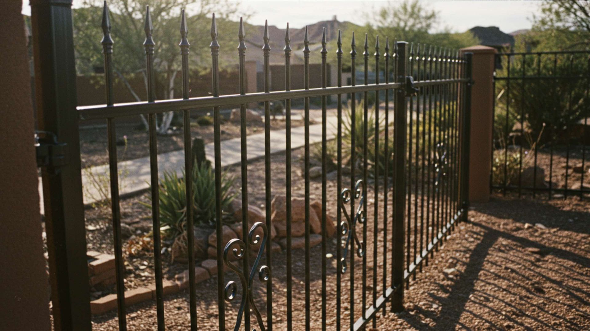 Black metal picket fence with pointed finials enclosing a desert landscape with gravel and native plants.