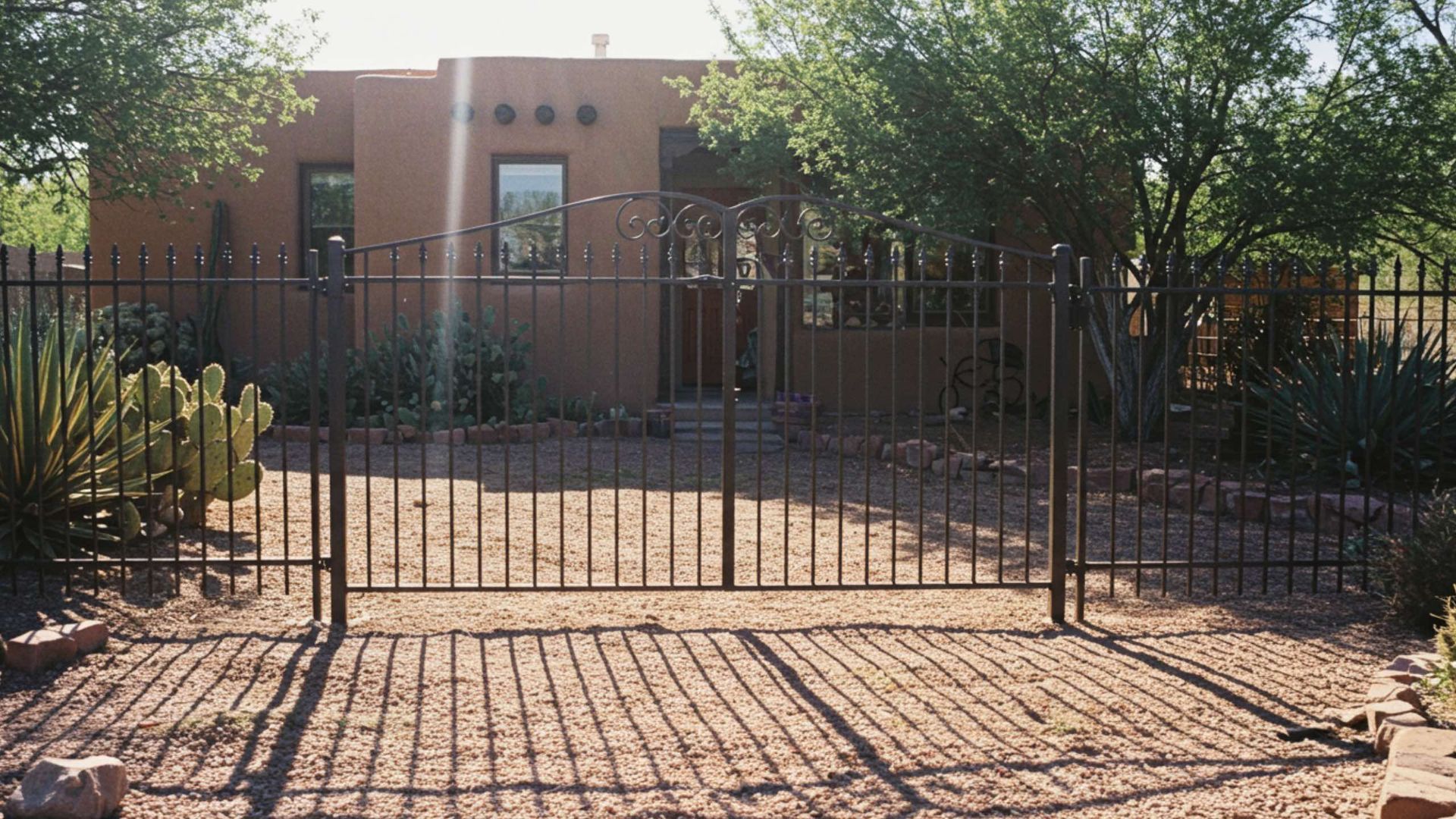A tan adobe house viewed behind a dark metal fence and gate in a sunny, desert-landscaped yard.