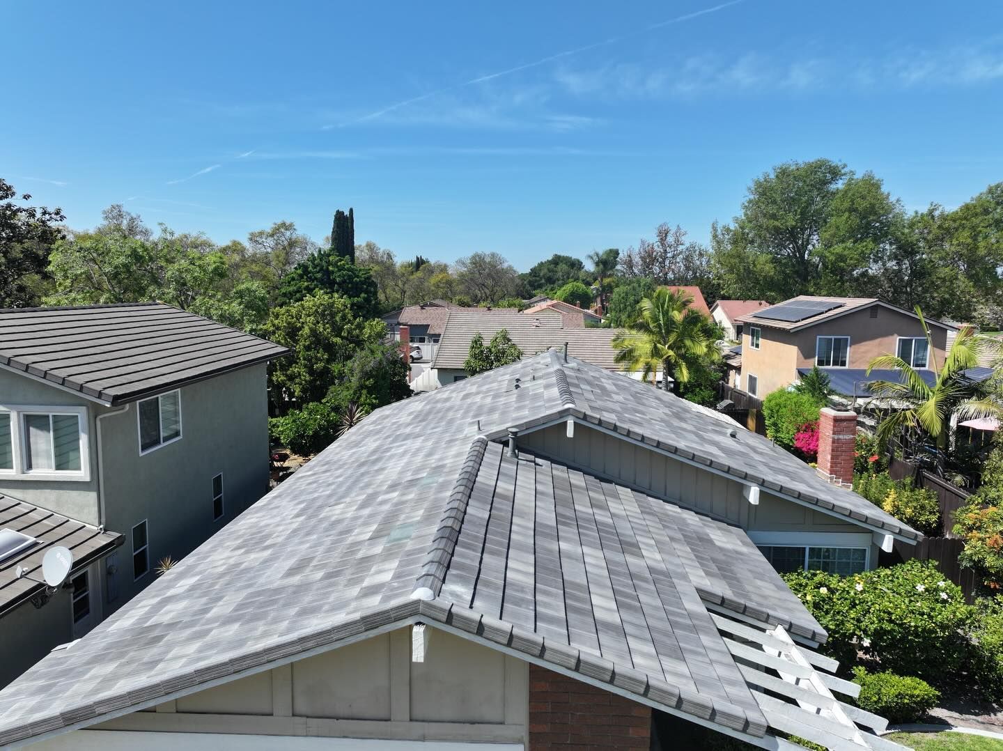 An aerial view of a gray tiled residential roof on a sunny day, surrounded by trees and neighboring houses.