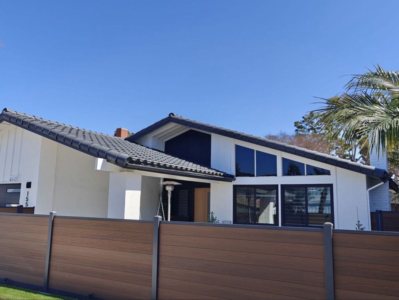 A white mid-century modern home with dark trim and roof, set behind a horizontal wooden privacy fence under a clear sky.