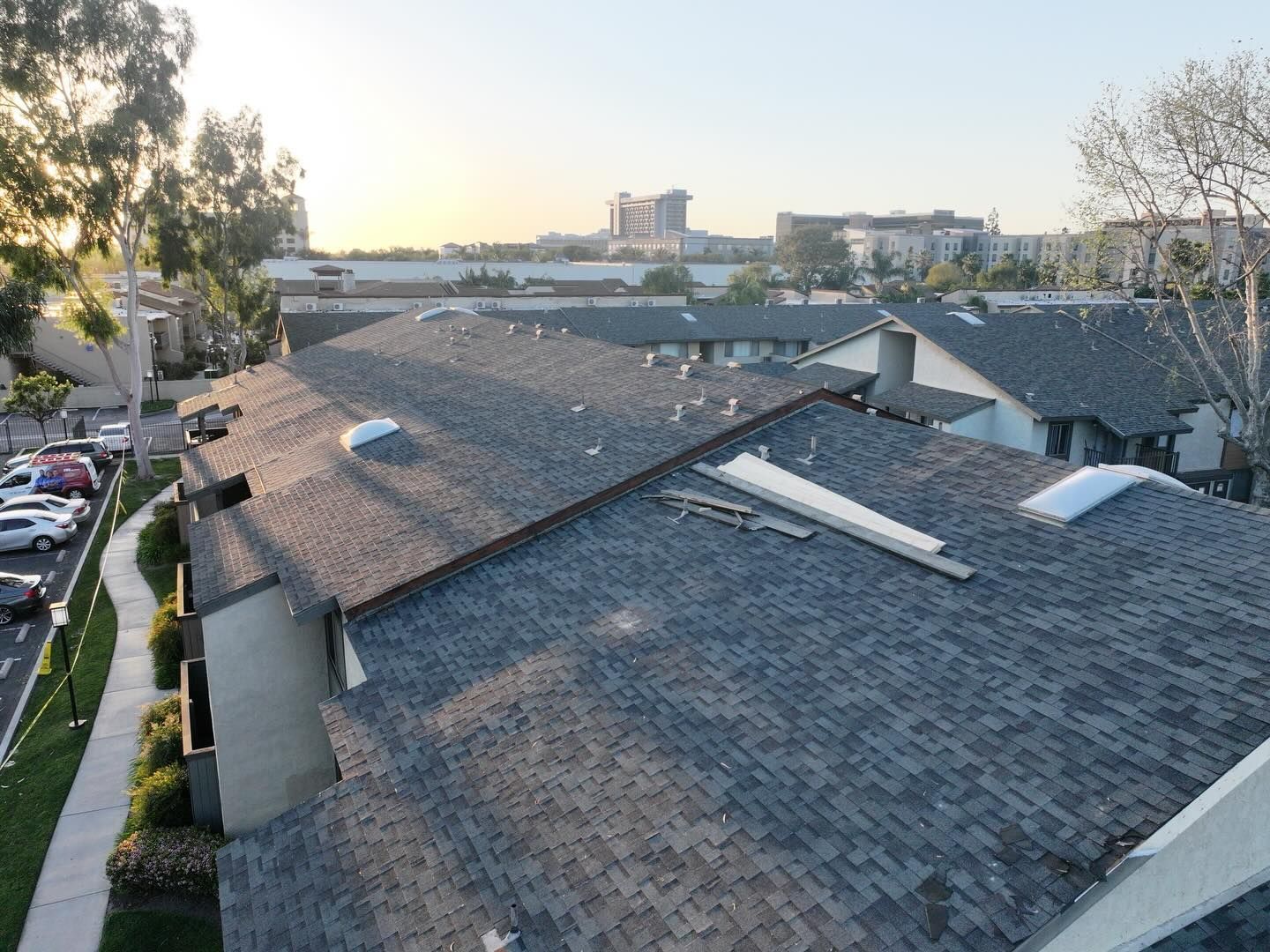 A high-angle view of a gray asphalt shingle roof with skylights, featuring nearby trees, a parking lot, and city buildings.