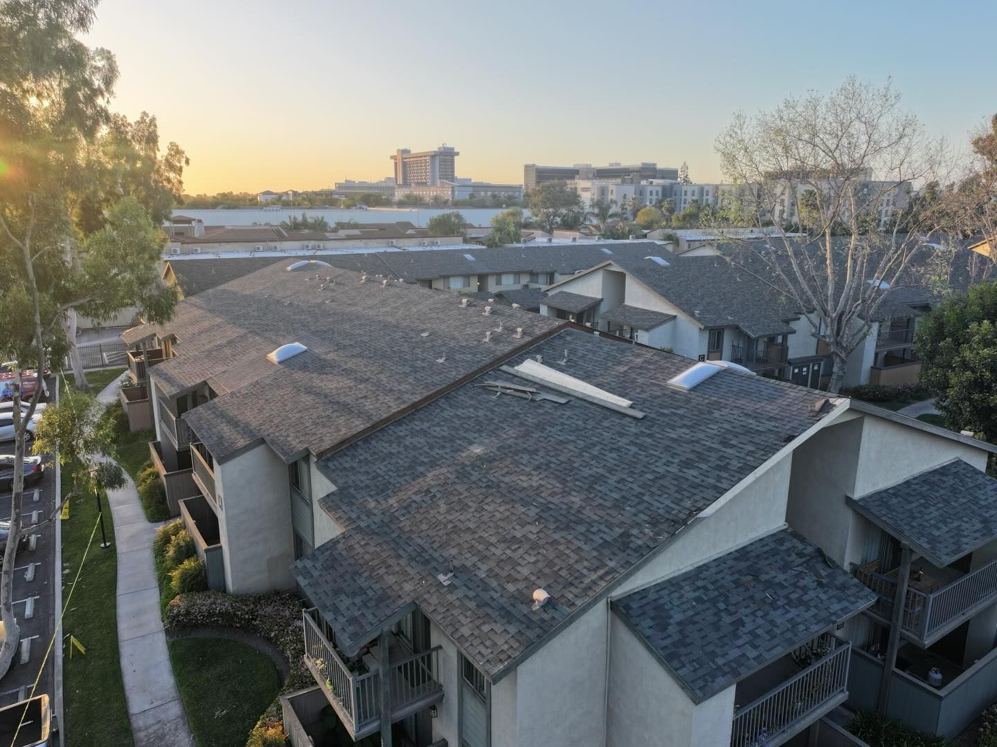 An elevated view of apartment buildings with gray shingled roofs at sunset, with distant buildings in the background.