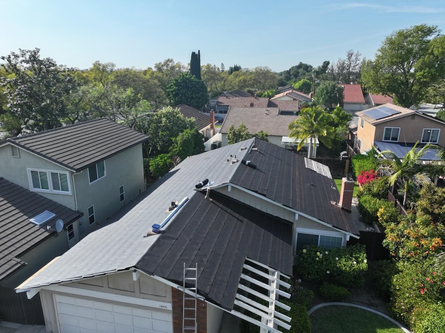 An aerial view shows a residential roof being replaced with dark shingles, with a ladder leaning against the side.