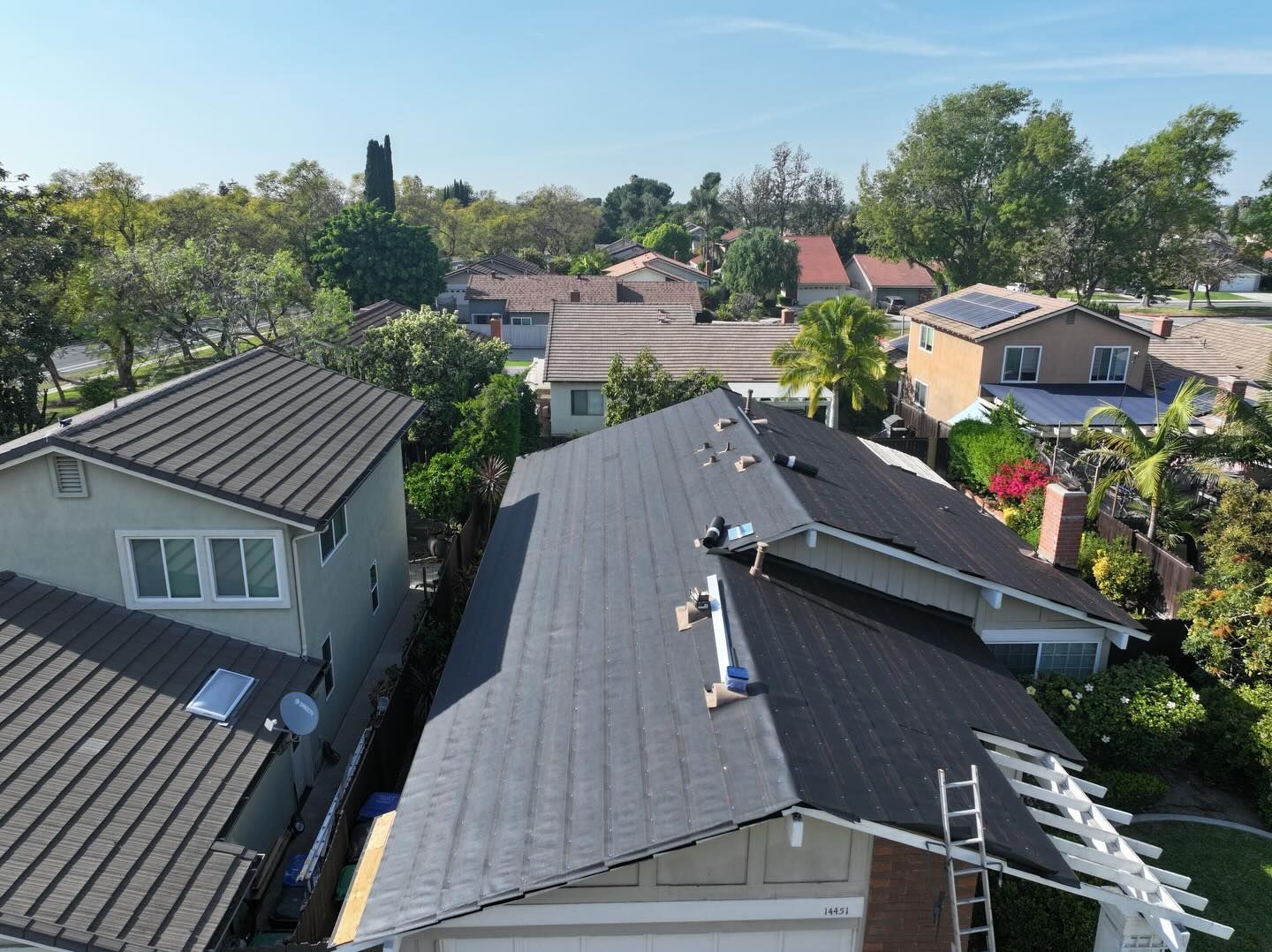 High-angle view of several suburban houses with dark shingled roofs on a sunny day, including a ladder leaned against one.