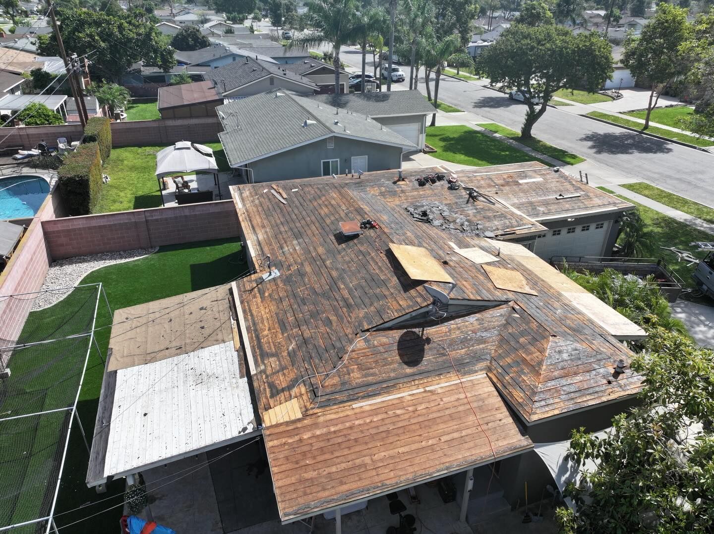 An aerial view of a suburban house during roof repairs, showing exposed wooden decking and patches of new roofing material.