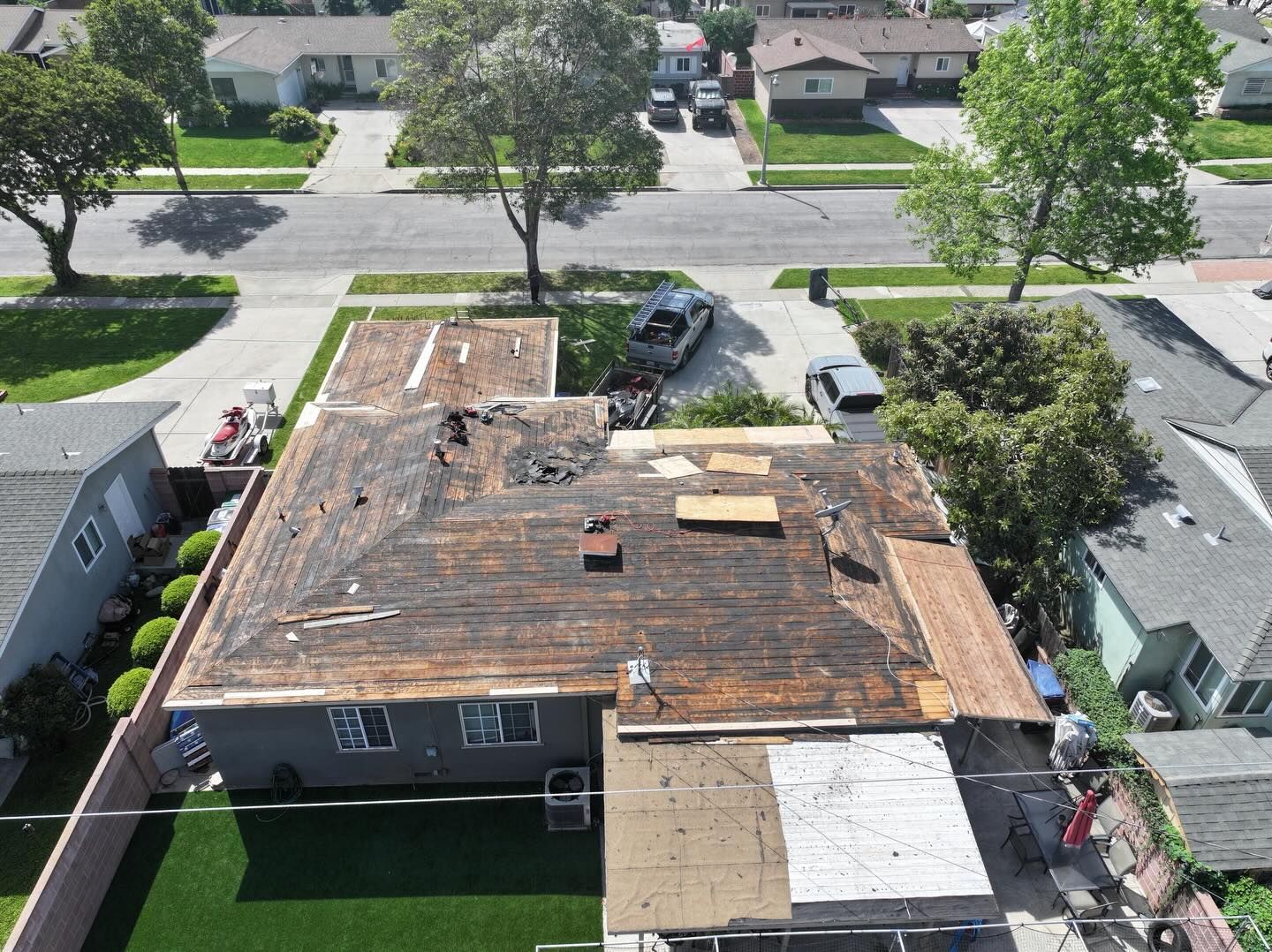 An aerial view of a house during roof replacement, showing bare plywood decking, construction materials, and a dumpster.