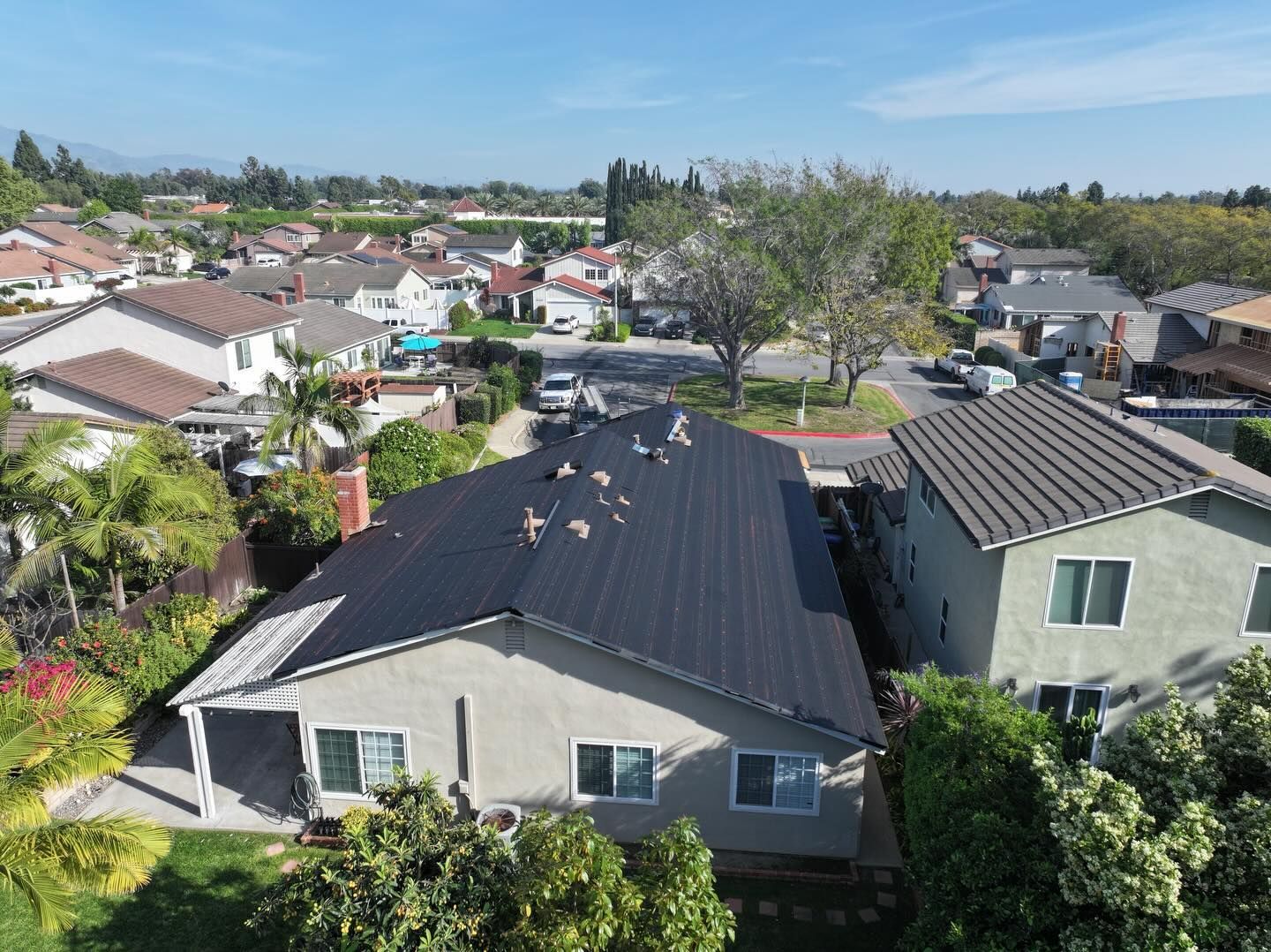 An aerial view of a single-story suburban house with a dark metal roof, surrounded by trees and neighboring homes.