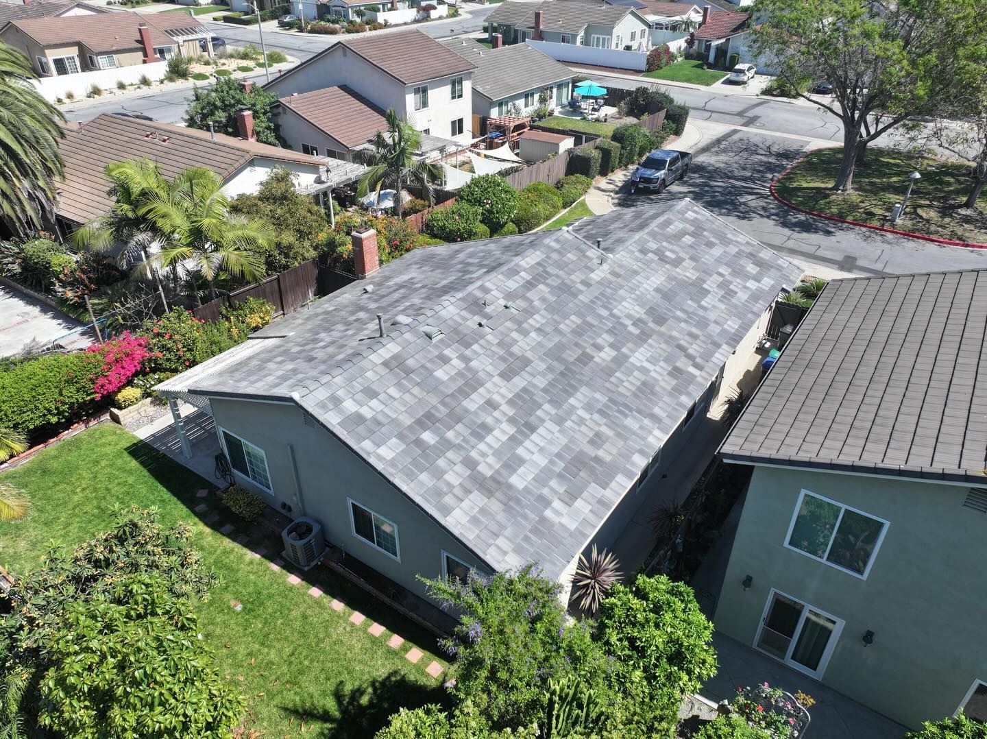 An aerial view of a gray shingled roof on a suburban house next to another home, surrounded by lawns and street trees.