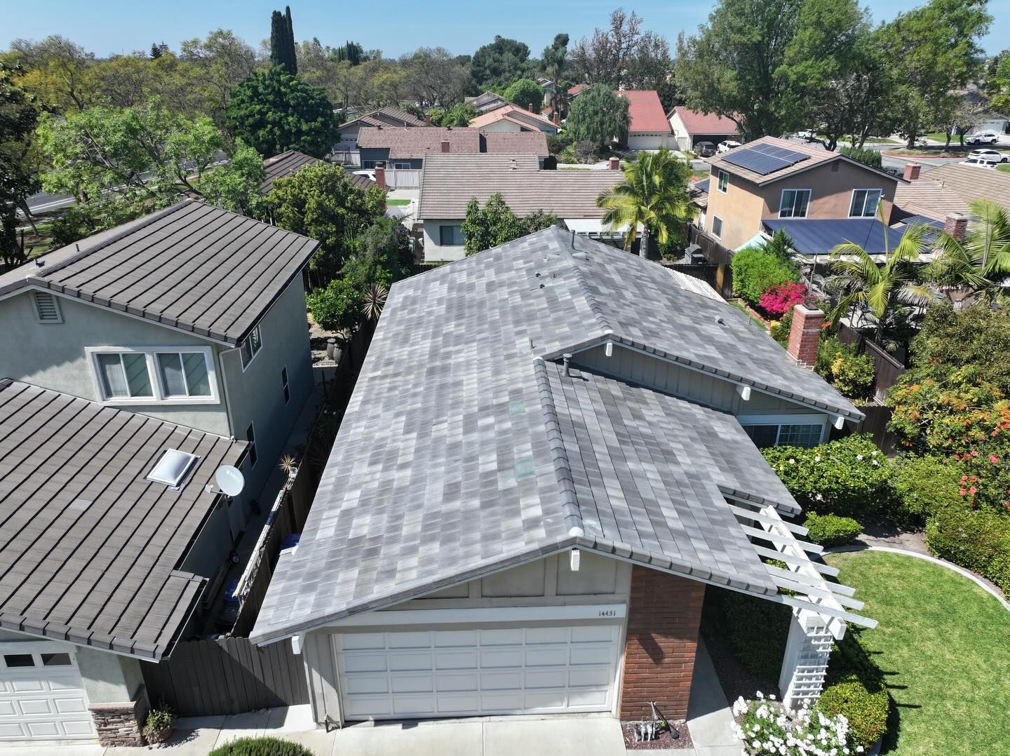 An aerial view of a residential street with grey-roofed houses, trees, and sunny blue skies.