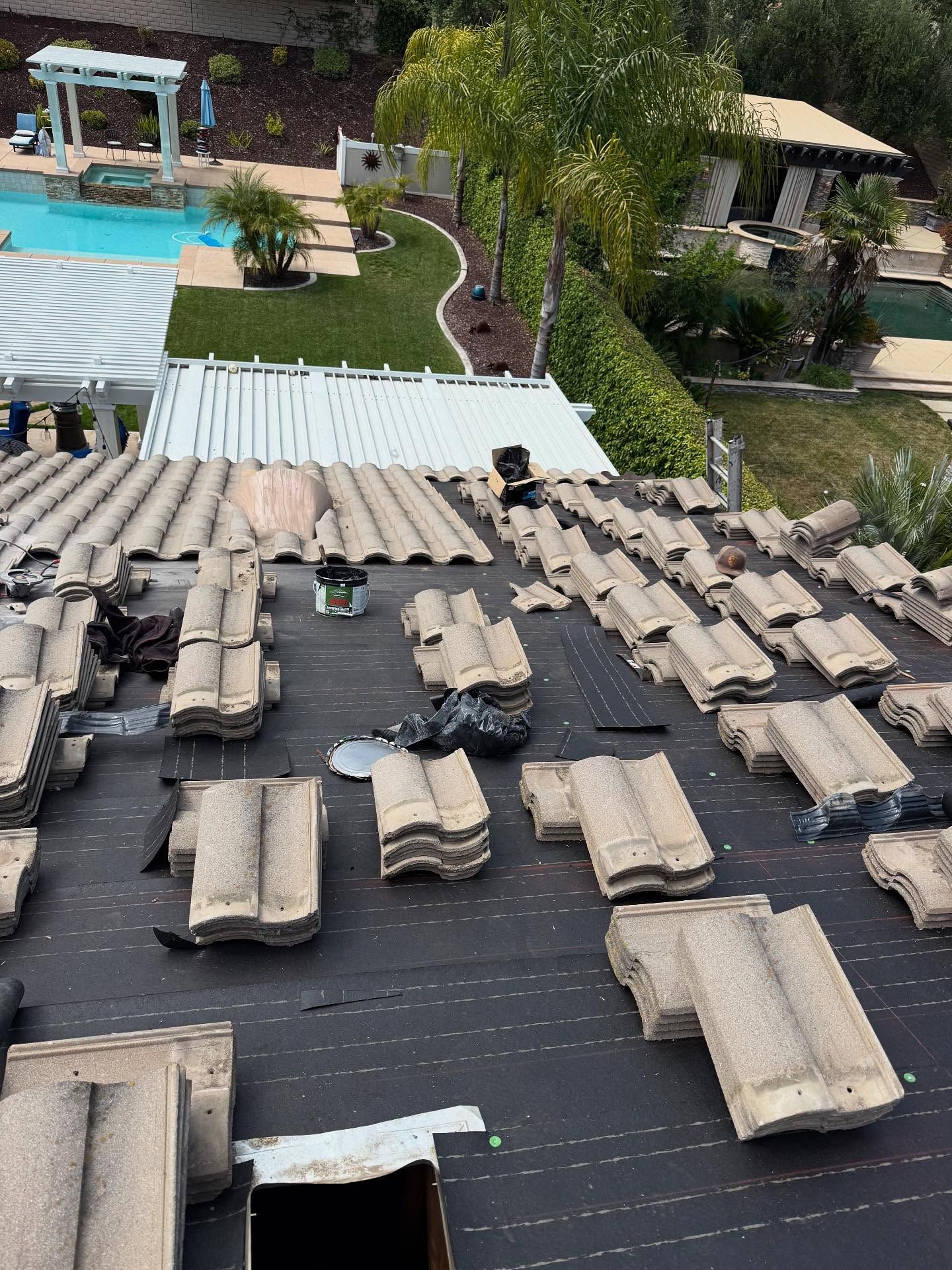 A residential roof undergoing repairs with loose clay tiles scattered across the black underlayment, overlooking a pool.