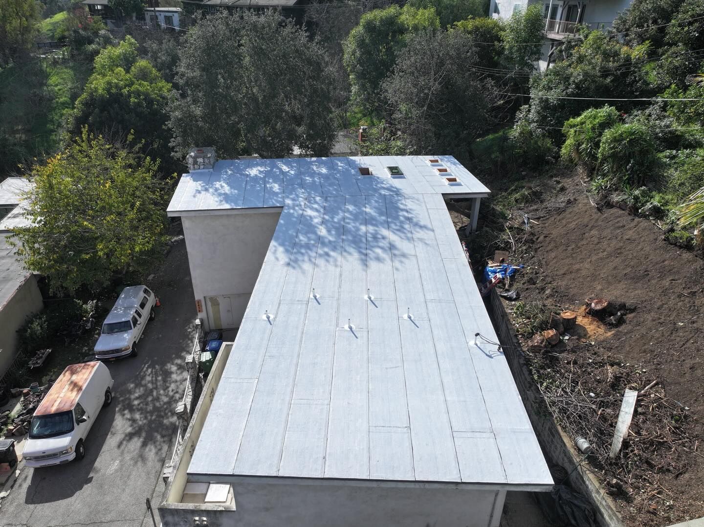 An aerial view of a flat, light-gray roof on a building, flanked by a driveway with two white vehicles and a hillside.
