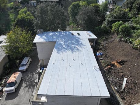 An aerial view of a flat, light-gray roof on a building, flanked by a driveway with two white vehicles and a hillside.