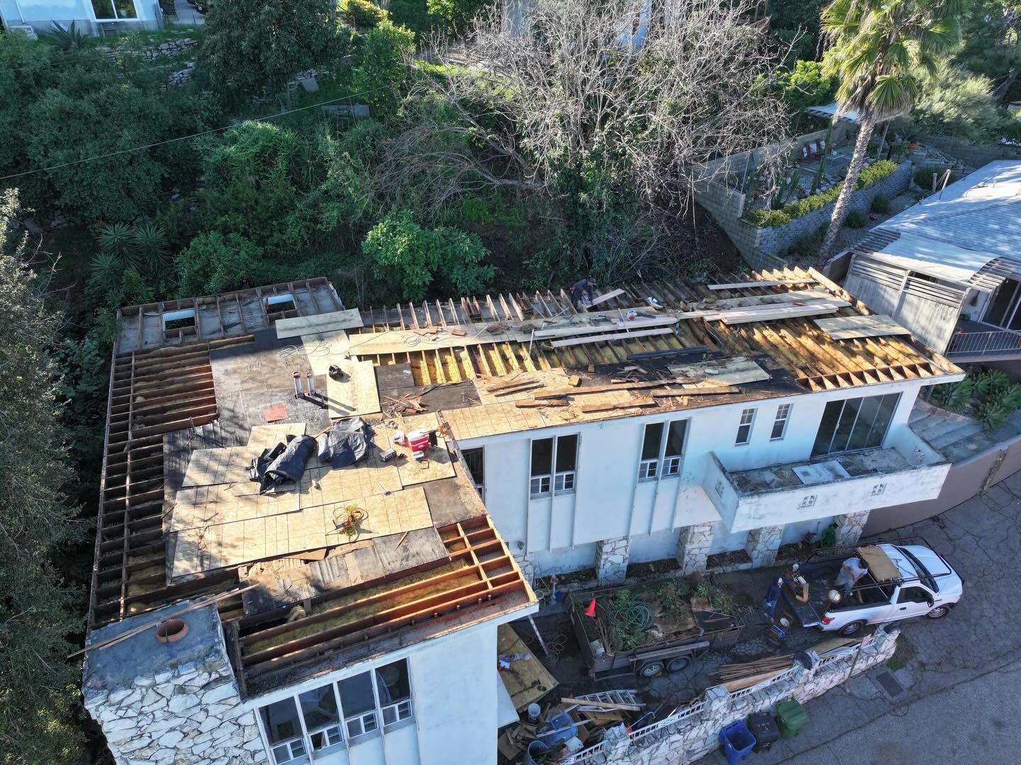 Aerial view of a white, multi-level house with its roof under construction, surrounded by trees and a parked white truck.
