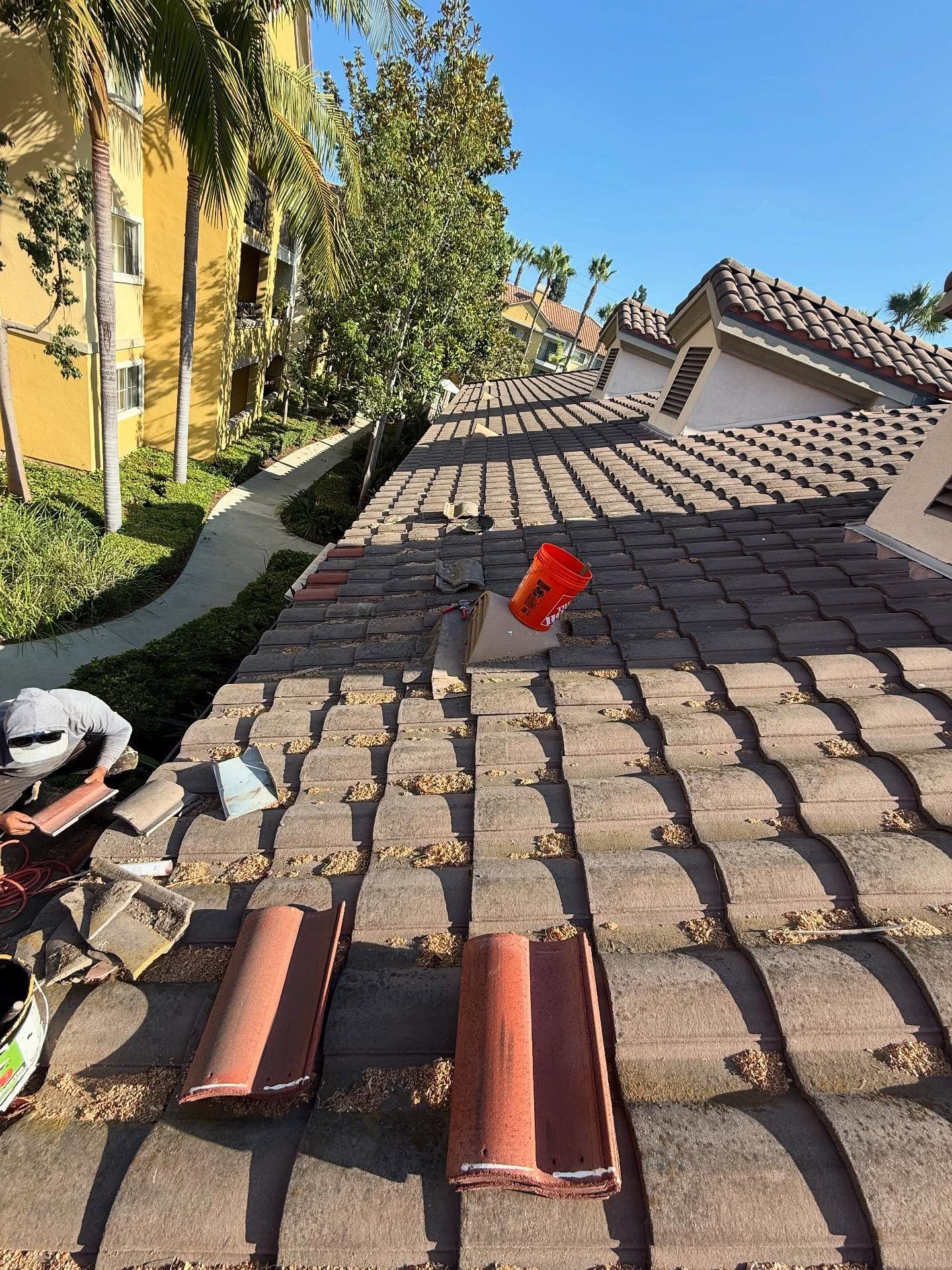 A worker performs roof repairs on a tiled roof, with spare tiles and a bucket resting on the slope.