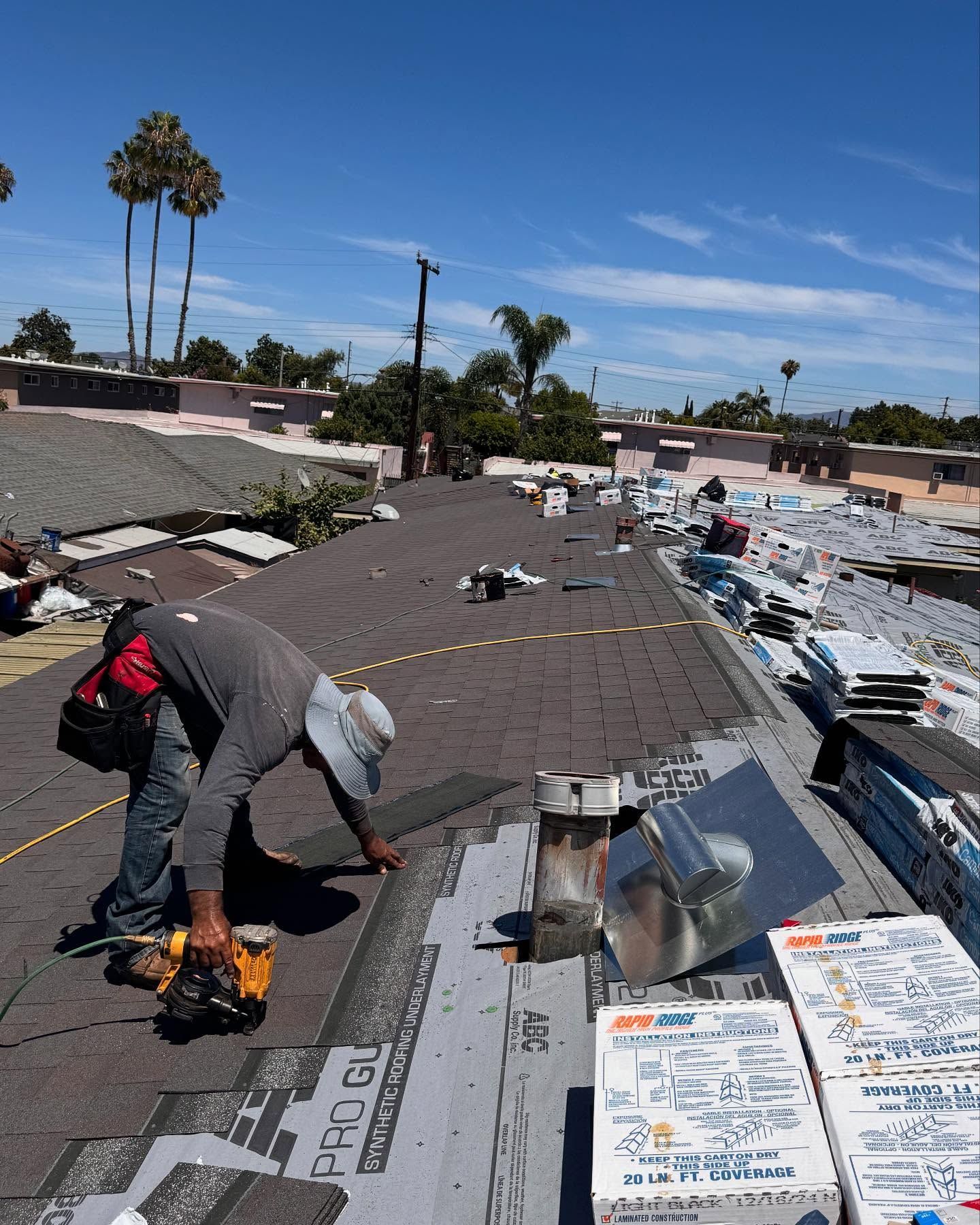 A worker wearing a hat and tool belt installs shingles on a sloped residential roof on a sunny day.