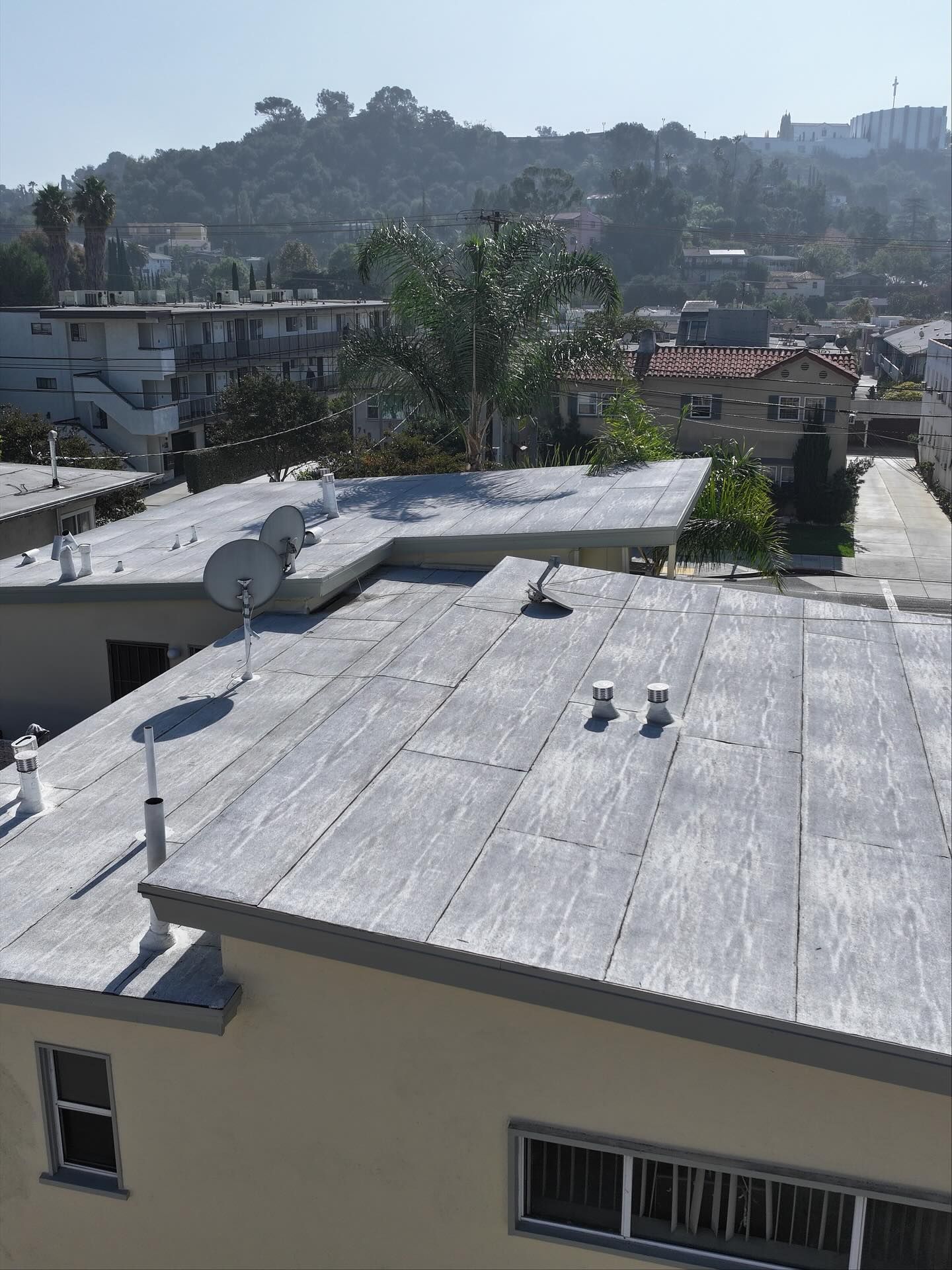 An elevated, sunlit view of a flat, gray-coated residential roof with satellite dishes and vents, overlooking a neighborhood.