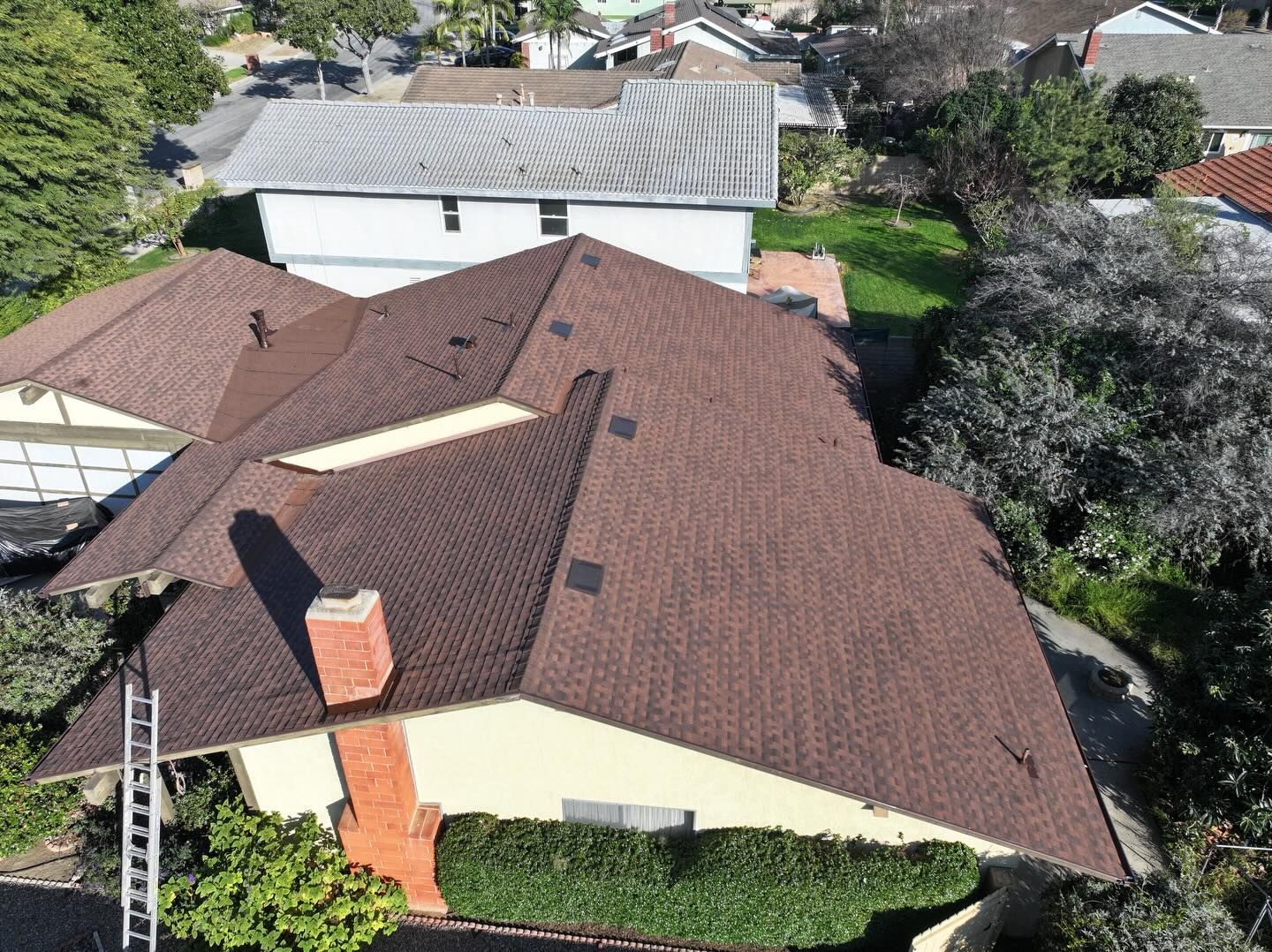 An aerial view of a house with a brown shingled roof, a brick chimney, and a ladder leaning against the side.