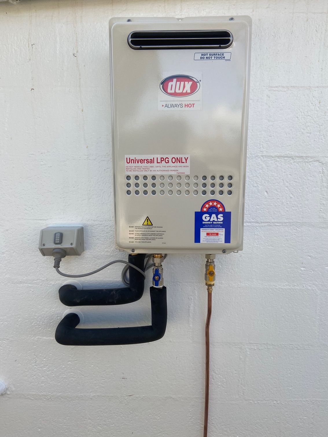 Wall-mounted Dux gas hot water heater, beige, with black pipes and copper gas line, against a white wall.
