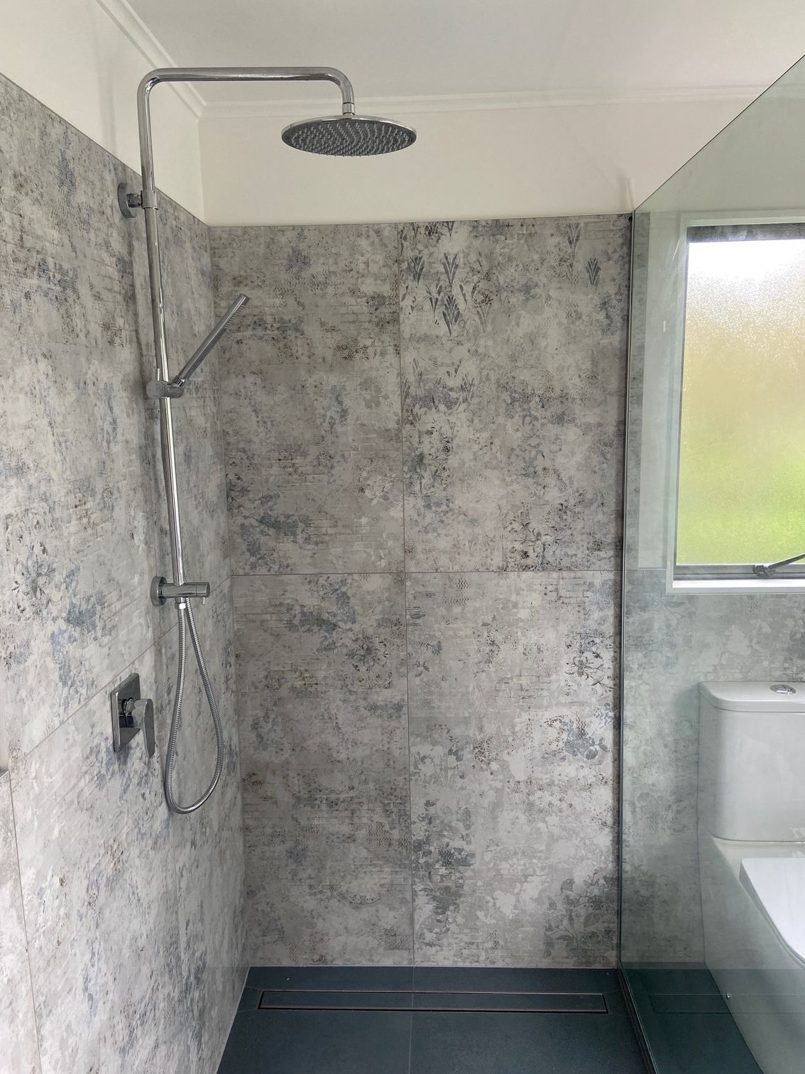 Shower with gray textured tile walls, rain shower head, and hand shower.