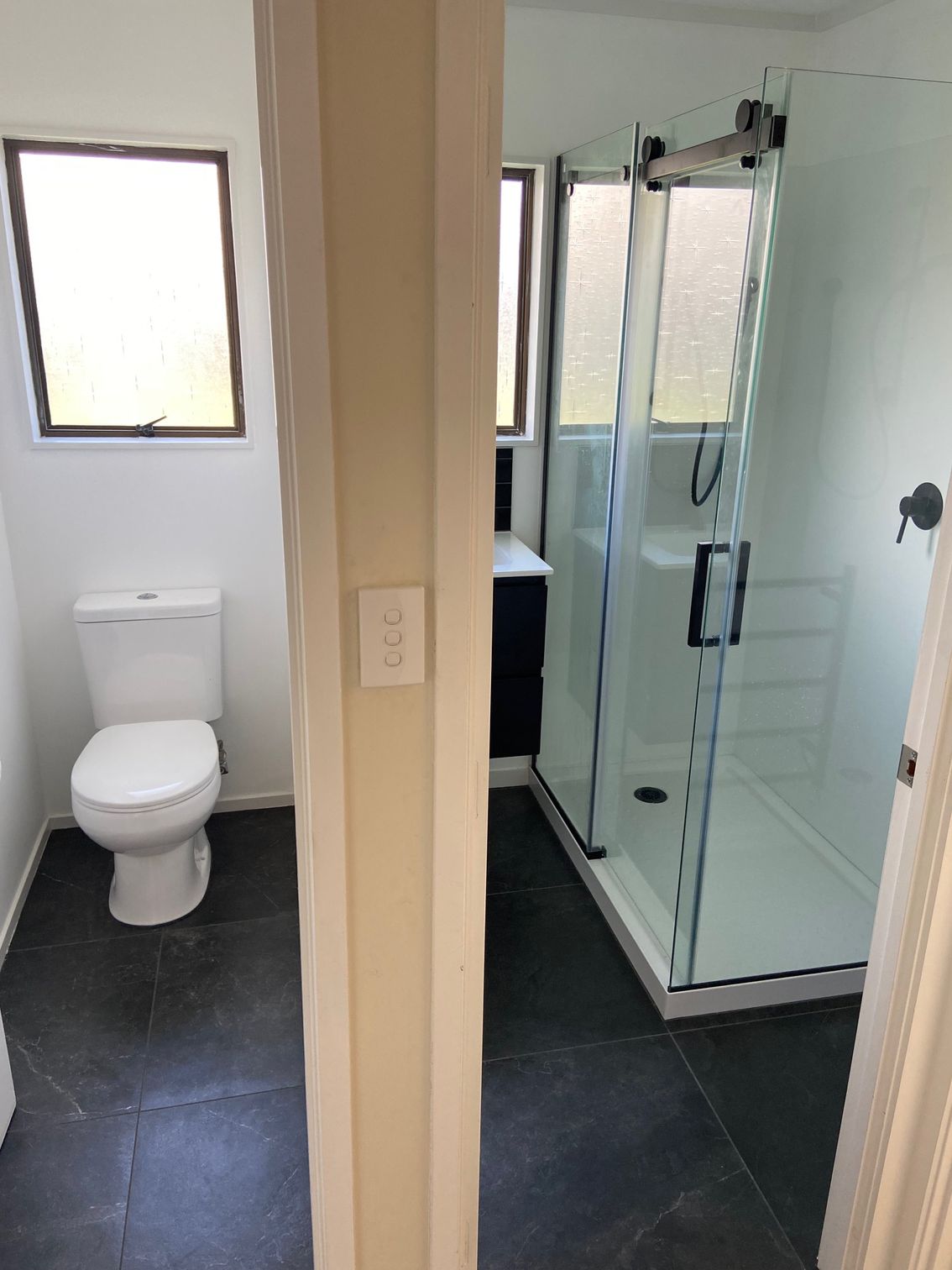 Bathroom with a toilet, shower, and vanity. Black tile floor, white walls, and a glass shower.