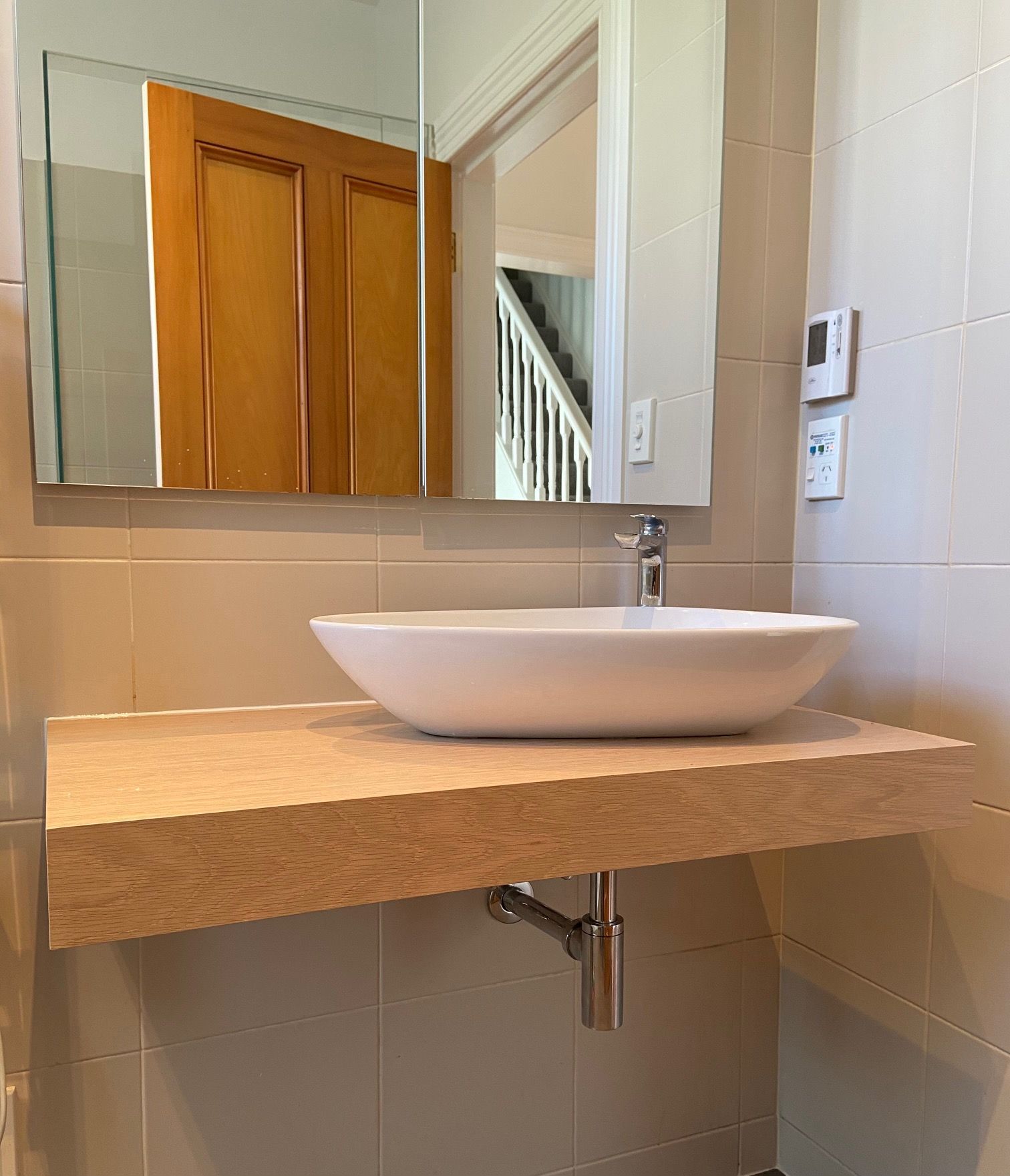 White vessel sink on a light beige floating countertop. Mirror reflects a door and staircase.