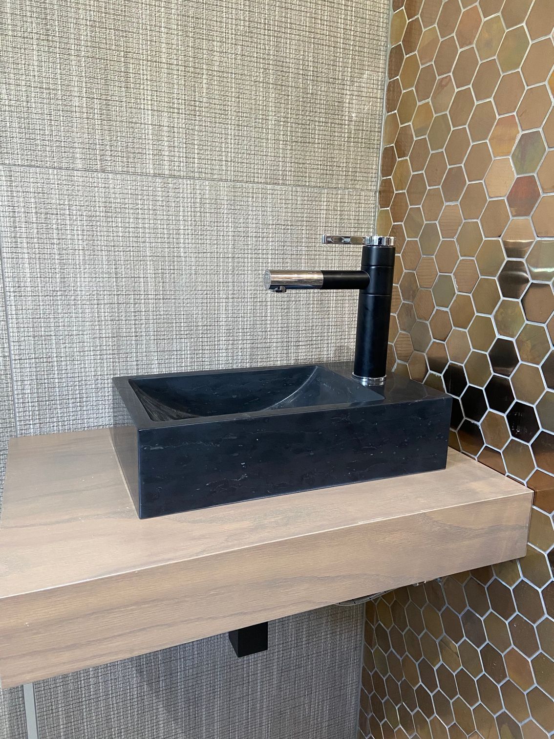 Black sink on wooden shelf, black faucet. Honeycomb tile wall, textured wallpaper.