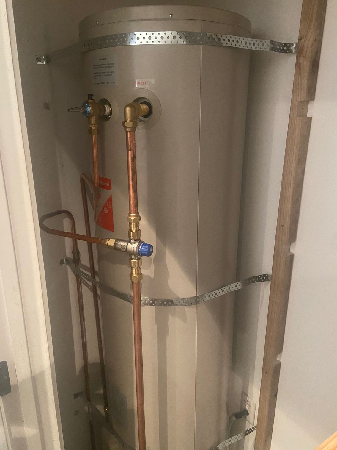 A tall, cylindrical water heater in a closet, with copper pipes and metal bands.