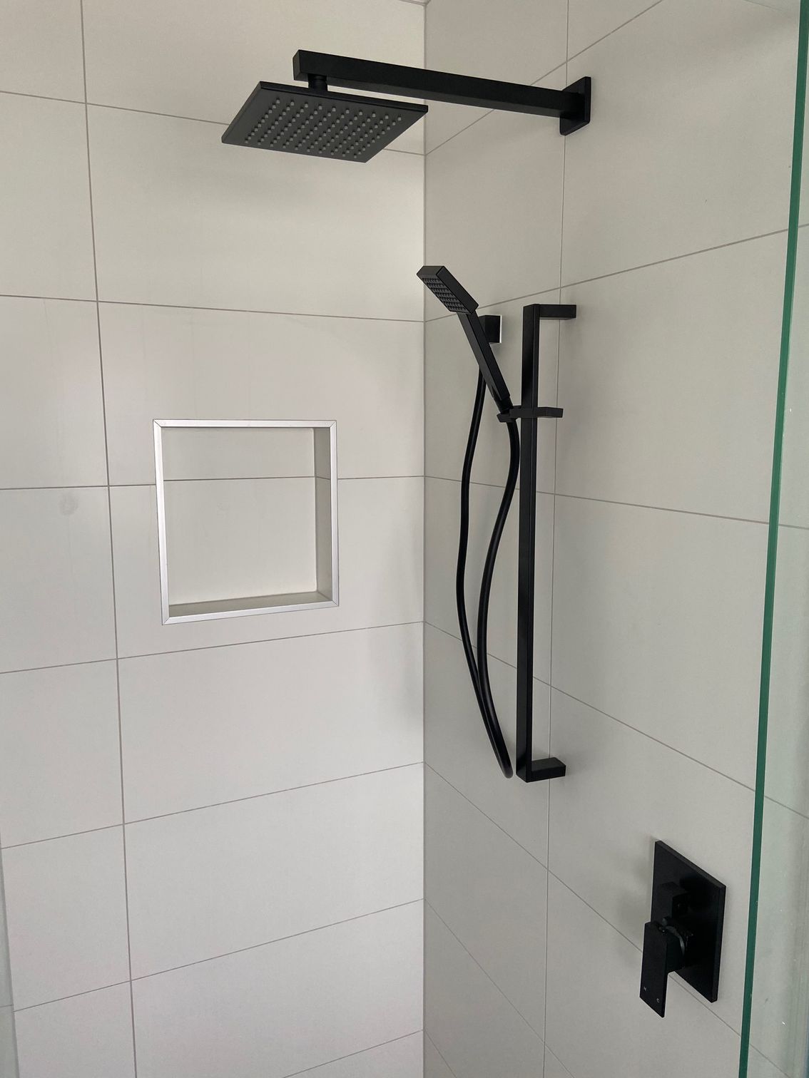 Black shower fixtures on white tiled wall; includes overhead showerhead, handheld sprayer, and controls.