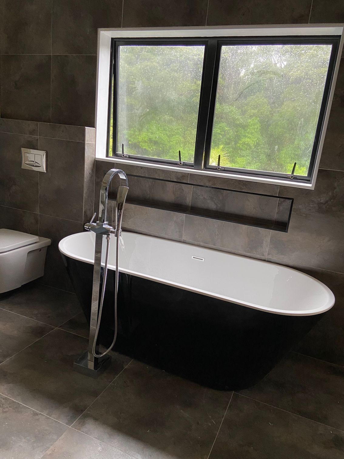 Bathroom with a black and white oval bathtub, a window, and gray tiled walls.
