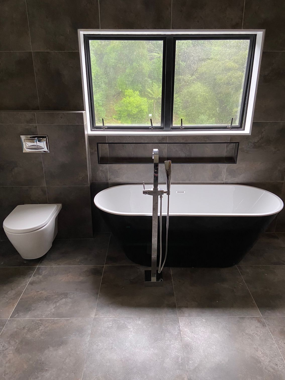 Bathroom with black bathtub, toilet, and window overlooking greenery. Dark gray walls and floor.