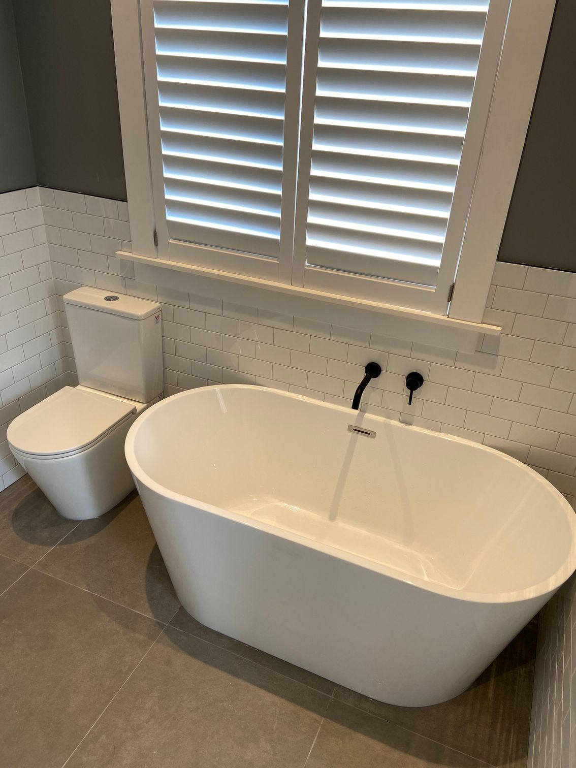 Bathroom with white tub, toilet, and window shutters. Grey floor and wall.