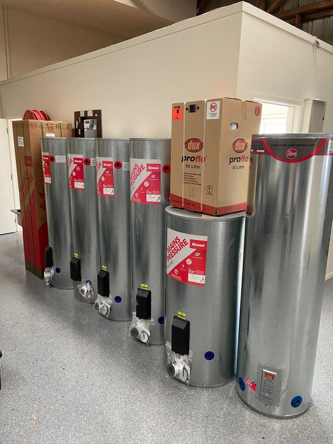 Row of new, silver water heaters in a room with white walls. Some have red labels and boxes on top.